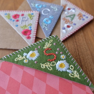 May include: Close-up of several handmade felt corner bookmarks. Each bookmark features embroidered floral designs and letters, with a variety of colors including green, pink, and blue. The bookmarks are placed on top of brown paper envelopes.