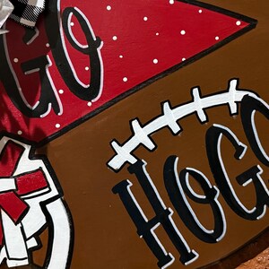 May include: A wooden football-shaped sign with the words "GO HOGS" in black and white. The sign has a red section with white polka dots and a white and red bow.