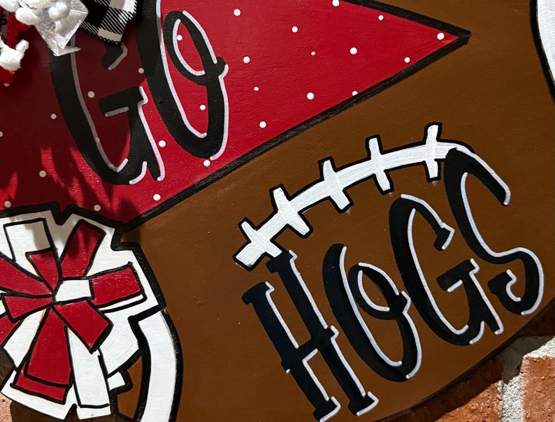 May include: A wooden football-shaped sign with the words "GO HOGS" in black and white. The sign has a red section with white polka dots and a white and red bow.