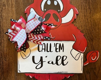 May include: A red wooden cutout of a cartoon wild boar holding a white rectangular sign with the black text CALL'EM Y'ALL. The boar features white tusks and a large decorative bow made of red, white, and black patterned ribbons attached to its side.