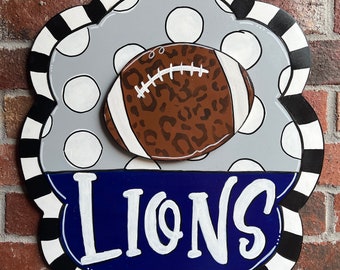 May include: A decorative wooden wall sign featuring a scalloped edge with black and white stripes. The top section is gray with white polka dots and a brown leopard print football. The bottom section is blue with the white text LIONS.