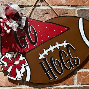 May include: A brown football-shaped wooden sign with the words "GO HOGS" in black script. The sign is decorated with a red and white bow and pom-poms. A rope is attached for hanging.