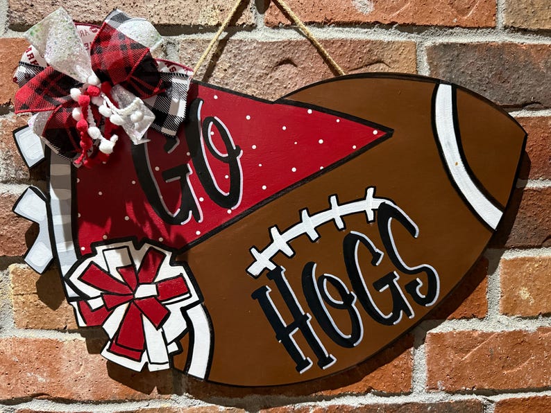 May include: A brown football-shaped wooden sign with the words "GO HOGS" in black script. The sign is decorated with a red and white bow and pom-poms. A rope is attached for hanging.