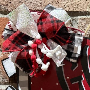 May include: A decorative bow with red and black plaid ribbon, white glitter ribbon, and red and white pom-poms. The bow is attached to a red surface with white polka dots and black accents. The bow has a rustic, festive appearance.
