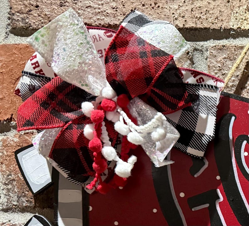 May include: A decorative bow with red and black plaid ribbon, white glitter ribbon, and red and white pom-poms. The bow is attached to a red surface with white polka dots and black accents. The bow has a rustic, festive appearance.