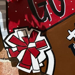 May include: A wooden football-shaped sign with the words "GO" and "HOME" in black and white. The sign has a red and white polka-dotted section and a brown section resembling a football. A red bow is also visible.
