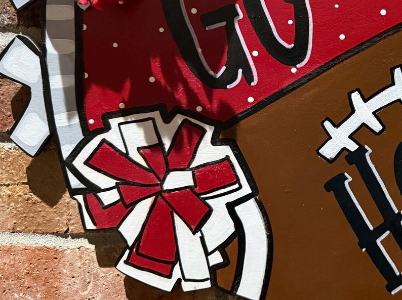 May include: A wooden football-shaped sign with the words "GO" and "HOME" in black and white. The sign has a red and white polka-dotted section and a brown section resembling a football. A red bow is also visible.
