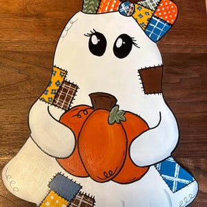 May include: A white ghost cutout featuring a large orange pumpkin held in its arms. The ghost is decorated with various colorful patchwork patterns in blue, yellow, orange, and brown, and wears a matching patchwork bow on its head.