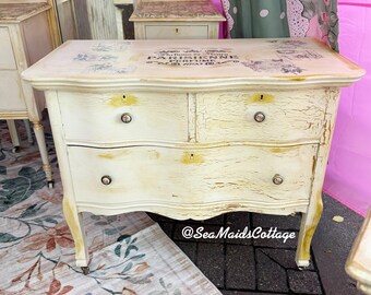May include: A distressed, off-white painted chest of drawers with two drawers and a top drawer. The chest has a curved front and legs, with a decorative floral design on the top. The text "PARISIENNE PERFUME" is printed on the top.