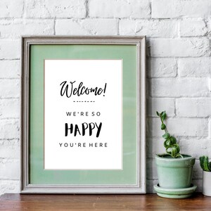 May include: A framed sign with a green mat and white background. The sign says "Welcome! We're so happy you're here". The sign is sitting on a wooden table in front of a white brick wall.