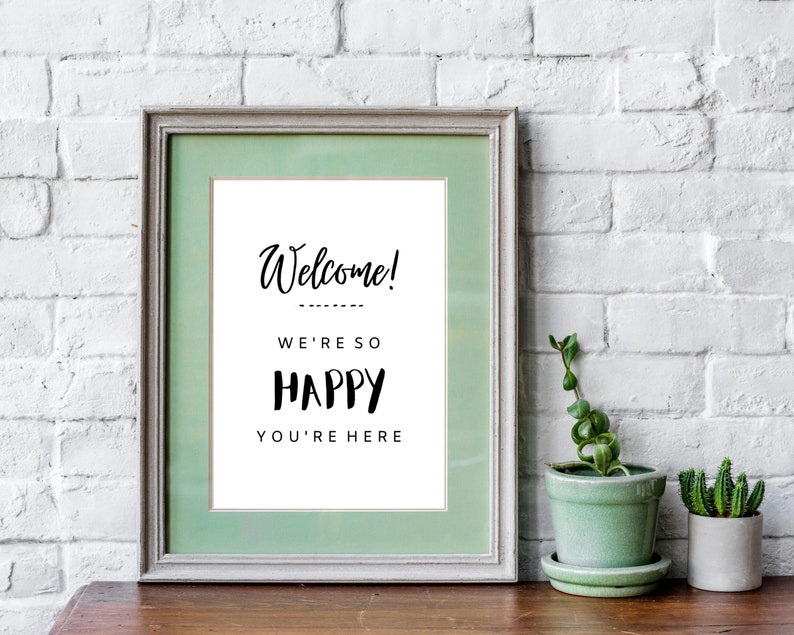 May include: A framed sign with a green mat and white background. The sign says "Welcome! We're so happy you're here". The sign is sitting on a wooden table in front of a white brick wall.