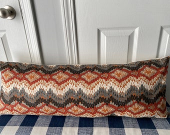 Op de afbeelding: Langwerpig, rechthoekig kussen met een geometrisch patroon in bruin, oranje, grijs en wit. Het kussen ligt op een blauw-wit geruit oppervlak. Ideaal voor een bank of bed.