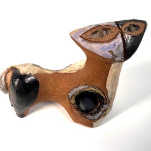 May include: A ceramic sculpture of a stylized dog with brown, black, white, and blue glaze. The dog is in a playful pose with its head tilted up.