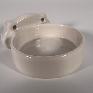 May include: Off-white ceramic pet bowl with a rounded, open design. The bowl has a mounting bracket with two holes for secure placement. The bowl is designed for pets and is made of ceramic.