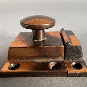 May include: A vintage copper and black door latch with a round knob. The latch has two holes on each side and a rectangular shape.
