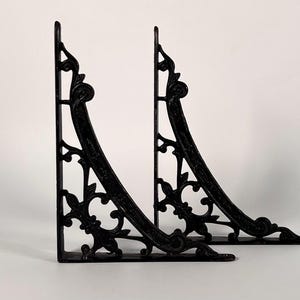 May include: Two black, ornate shelf brackets. The brackets are made of cast iron and feature a decorative, curved design with scrollwork and floral elements. The brackets are L-shaped and ready to be mounted on a wall.
