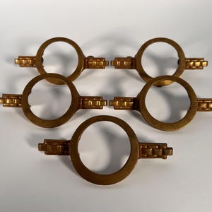May include: Five antique brass-toned cabinet pulls. Each pull features a circular handle with a rectangular decorative element on either side. The pulls are arranged on a white surface, showcasing their design and finish.