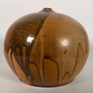 May include: A handcrafted, spherical ceramic vase with a small spout. The vase is divided into two sections, one a darker brown and the other a lighter tan. Dark brown drips cascade down the lighter side, creating a unique, textured appearance.