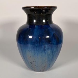 May include: A glossy, blue ceramic vase with a dark brown rim. The vase features a gradient of blues, transitioning from a deep navy at the top to a lighter, speckled blue at the base. The vase has a wide body and a slightly flared neck.