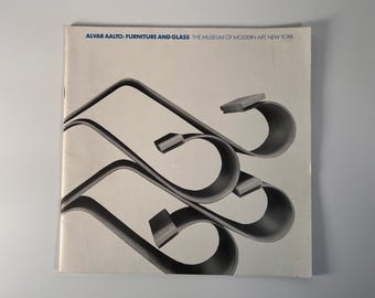 May include: A black and white photo of a book cover featuring the text "ALVAR AALTO: FURNITURE AND GLASS THE MUSEUM OF MODERN ART, NEW YORK" and a graphic design of three curved, overlapping shapes.