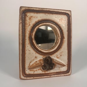 May include: A small, white ceramic mirror with a brown frame and a floral design. The mirror is round and has a reflective surface.
