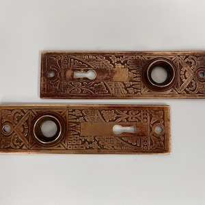 May include: Two antique brass door plates with intricate floral and geometric designs. Each plate has a keyhole and a round knob hole.