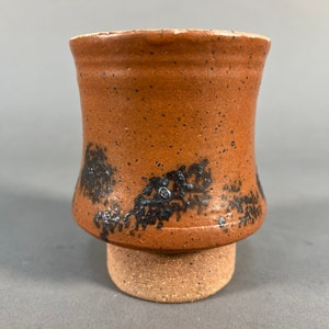 May include: A brown ceramic cup with a flared base and black speckles. The cup is sitting on a small, round, brown base.