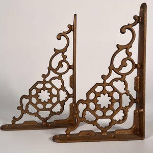 May include: Two ornate, bronze-colored shelf brackets. Each bracket features a decorative, openwork design with scrolling details and a geometric pattern. The brackets are made of metal and are designed to be mounted on a wall.