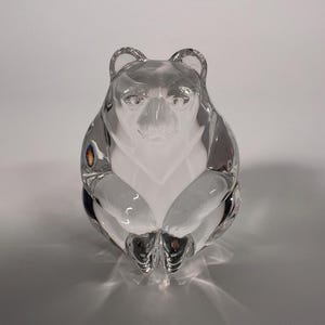 May include: A clear glass bear figurine with a seated pose. The bear has detailed features, including ears, eyes, and paws. The glass is transparent, allowing light to pass through, creating shadows. The figurine is a decorative item.