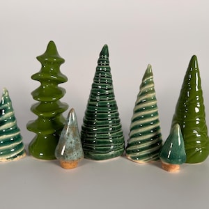 May include: A collection of ceramic Christmas trees in various shades of green. The trees have different shapes and textures, including spiral and stacked designs. Some trees have a glossy finish, while others have a matte appearance. The trees are arranged on a white surface.