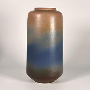 May include: A tall, cylindrical ceramic vase with a gradient color scheme. The top is a warm brown, transitioning through blue and green to a darker brown base. The vase has a textured, crackled surface.