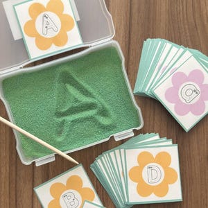 May include: A clear plastic container with green sand and the letter "A" imprinted. The set includes flower-shaped cards with letters A, B, C, and D, and a wooden stylus. The cards have a yellow flower design with a white center and a light green border.
