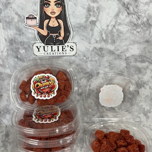 May include: Clear plastic containers filled with red-colored candies, stacked and arranged on a marble-patterned surface. The containers have labels that read "Chamoy Rim Dip." A sticker of a woman holding a cake and the text "Yulie's Creations" is also visible.