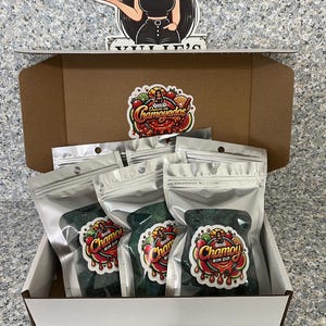 May include: A white box contains several sealed bags of Chamoy Rim Dip. Each bag has a colorful sticker with the product name and a graphic of a woman and fruit. A decorative cutout of a woman holding a cake is on top.