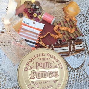May include: A vintage tin with the text "D. Politi & Sons Ltd. Politi's Fudge Est. 1870 London N.16."  The tin is surrounded by various craft supplies, including lace, ribbon, buttons, and thread.