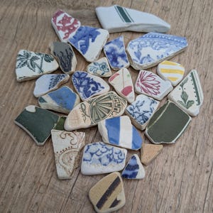 May include: A selection of ceramic fragments with diverse patterns and colours. The pieces display blue, green, red, and yellow tones, with floral and geometric designs. The shards are arranged on a wooden surface, highlighting their unique shapes and textures.