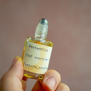 Puede incluir: Un pequeño frasco de vidrio con un aplicador de bola lleno de un líquido amarillo. La etiqueta dice "Protection 10ml conjure oil / apsara, perfumery".