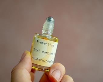 May include: A small glass bottle with a rollerball top filled with a yellow liquid. The label reads "Protection 10ml conjure oil / apsara, perfumery".