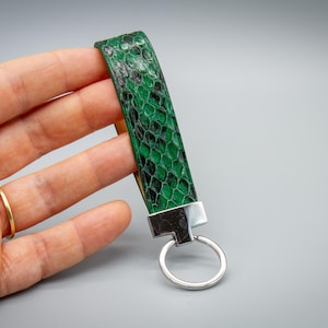 May include: A green and black snakeskin-patterned wrist strap keychain with a silver-colored metal clasp and key ring. The strap is held in a hand against a gray background.