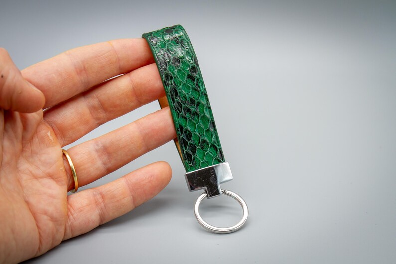 Peut inclure: Un porte-cl&eacute;s &agrave; dragonne avec un motif de peau de serpent vert et noir, avec un fermoir et un anneau porte-cl&eacute;s en m&eacute;tal argent&eacute;. La dragonne est tenue dans une main sur fond gris.