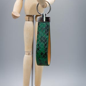 Peut inclure: Un porte-cl&eacute;s en cuir vert &agrave; motif peau de serpent, avec un anneau et des accents en m&eacute;tal argent&eacute;. Le porte-cl&eacute;s a une conception en boucle et un int&eacute;rieur contrastant de couleur beige. L'accessoire est pr&eacute;sent&eacute; sur un fond gris neutre.