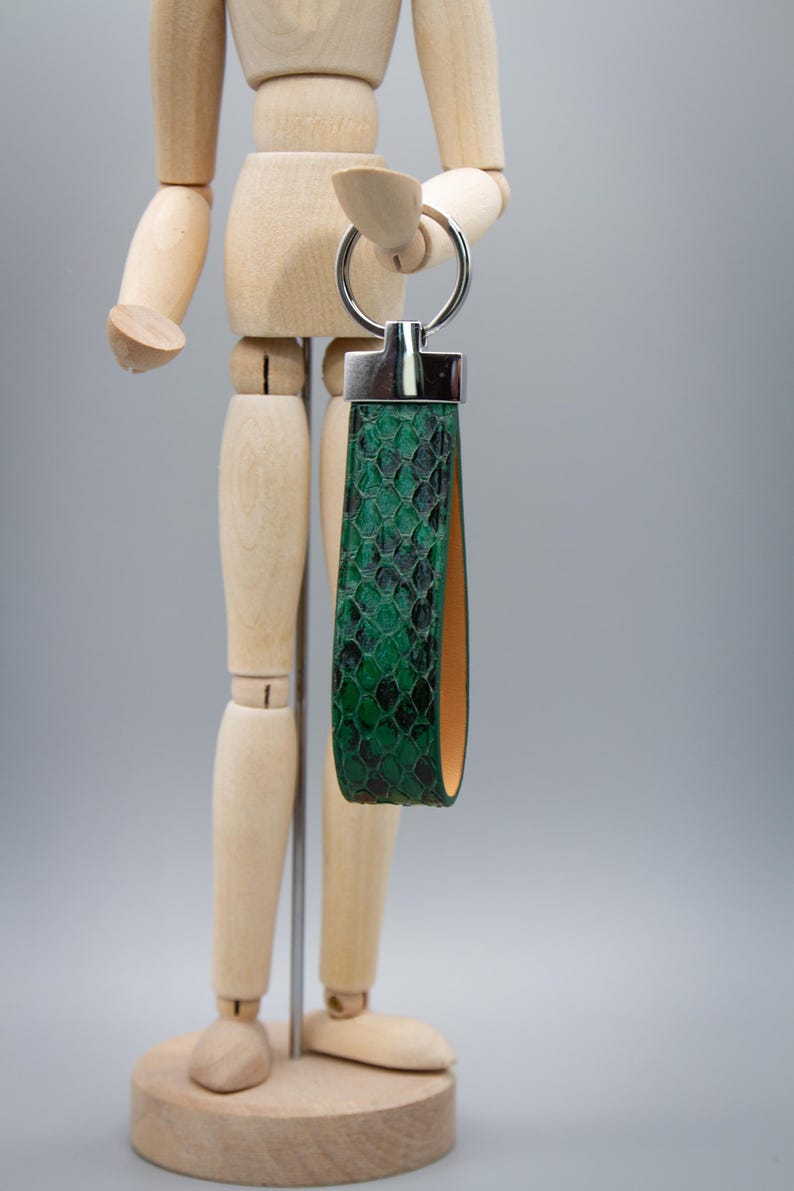 Peut inclure: Un porte-cl&eacute;s en cuir vert &agrave; motif peau de serpent, avec un anneau et des accents en m&eacute;tal argent&eacute;. Le porte-cl&eacute;s a une conception en boucle et un int&eacute;rieur contrastant de couleur beige. L'accessoire est pr&eacute;sent&eacute; sur un fond gris neutre.