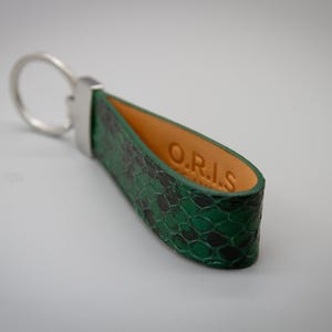 Peut inclure: Un porte-cl&eacute;s en cuir vert et noir &agrave; motif peau de serpent avec un anneau en m&eacute;tal argent&eacute;. L'int&eacute;rieur du porte-cl&eacute;s est en cuir beige, avec les lettres "O.R.I.S" estamp&eacute;es.