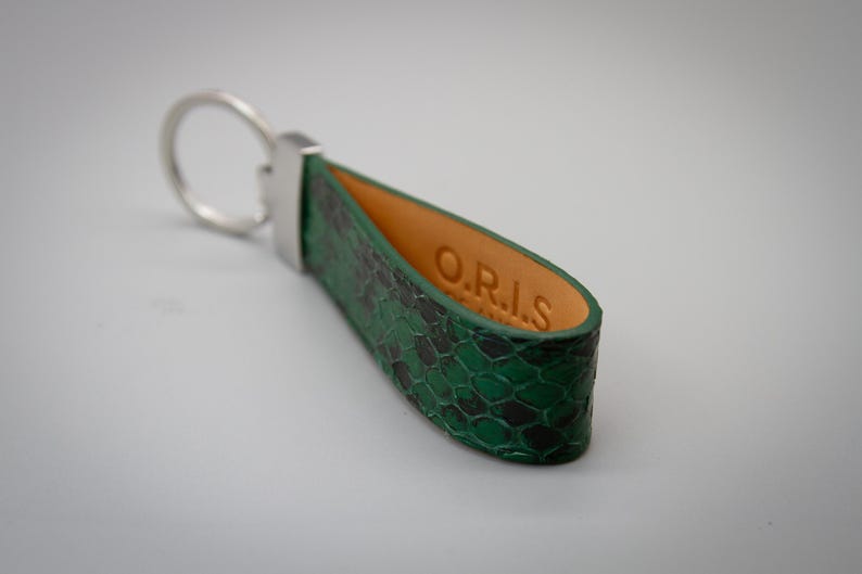 Peut inclure: Un porte-cl&eacute;s en cuir vert et noir &agrave; motif peau de serpent avec un anneau en m&eacute;tal argent&eacute;. L'int&eacute;rieur du porte-cl&eacute;s est en cuir beige, avec les lettres "O.R.I.S" estamp&eacute;es.