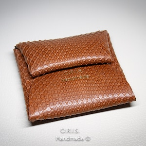 Pode incluir: Uma carteira de couro marrom com um padrão de pele de cobra. A carteira está dobrada ao meio e tem o texto "ORIS Handmade" gravado na frente.