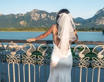 May include: A bride wearing a white wedding dress and veil stands on a bridge overlooking a lake and mountains in the distance.
