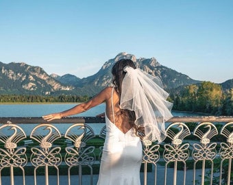 May include: A bride wearing a white wedding dress and veil stands on a bridge overlooking a lake and mountains. The bridge has an ornate metal railing.