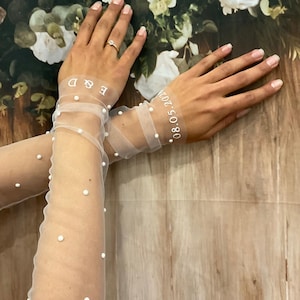 Könnte beinhalten: Transparente weiße Hochzeits-Handschuhe mit kleinen, gleichmäßig verteilten Perlen. Die Handschuhe zeigen die Initialen "E & D" und das Datum "08.05.2024" in Weiß. Der Hintergrund zeigt ein Blumenmuster und eine Holzstruktur.