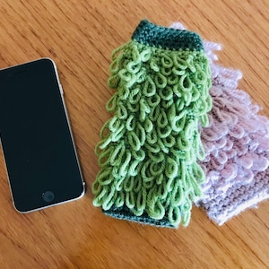 May include: A silver smartphone rests on a wooden surface next to two handmade phone cases. One case is green with looped yarn, the other is lavender. The cases are designed to protect the phone.