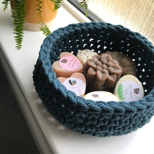 May include: A teal blue crocheted basket filled with various handmade soap bars. The soaps are in different shapes and sizes, including a heart, a flower, and a rectangle. The soaps are labeled with names like "Spa Clay Shampoo" and "Nale Marceli Soap".